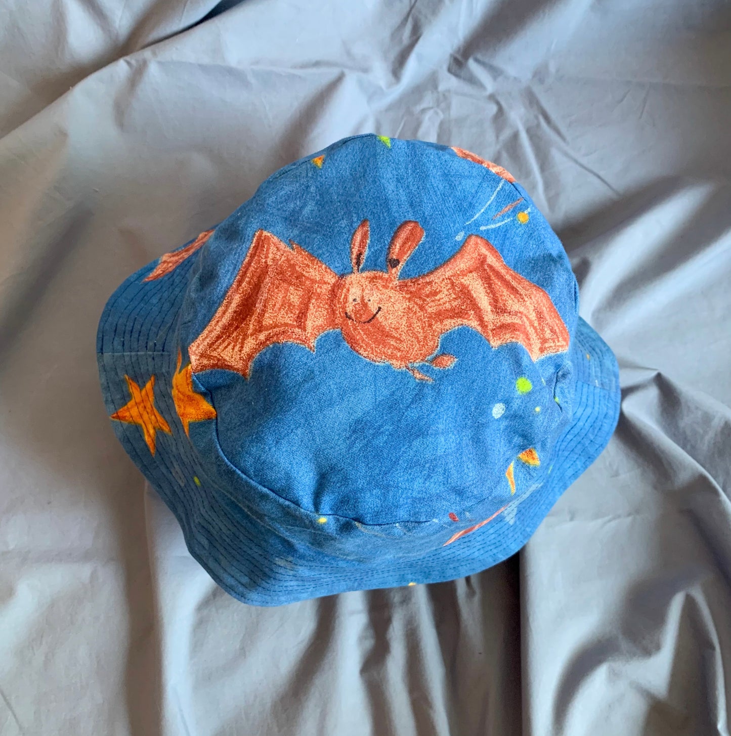 The top view of a blue bucket hat with a red cartoon bat and stars on the top, on a blue background