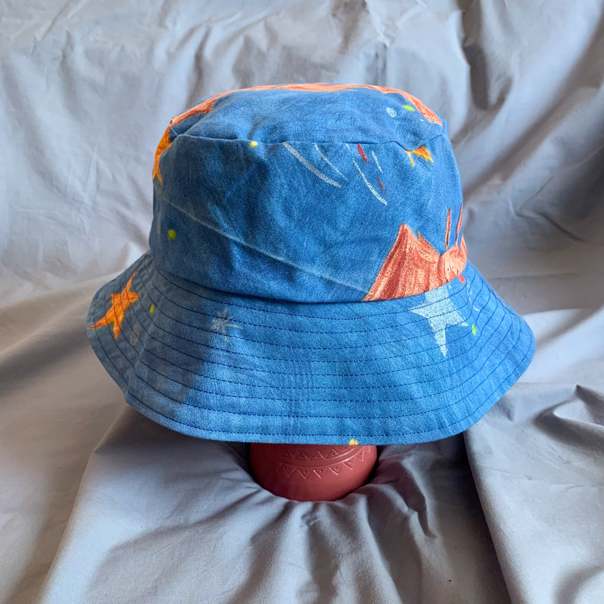 A blue bucket hat with part of a bat cartoon styled on a pink stone head against a blue background