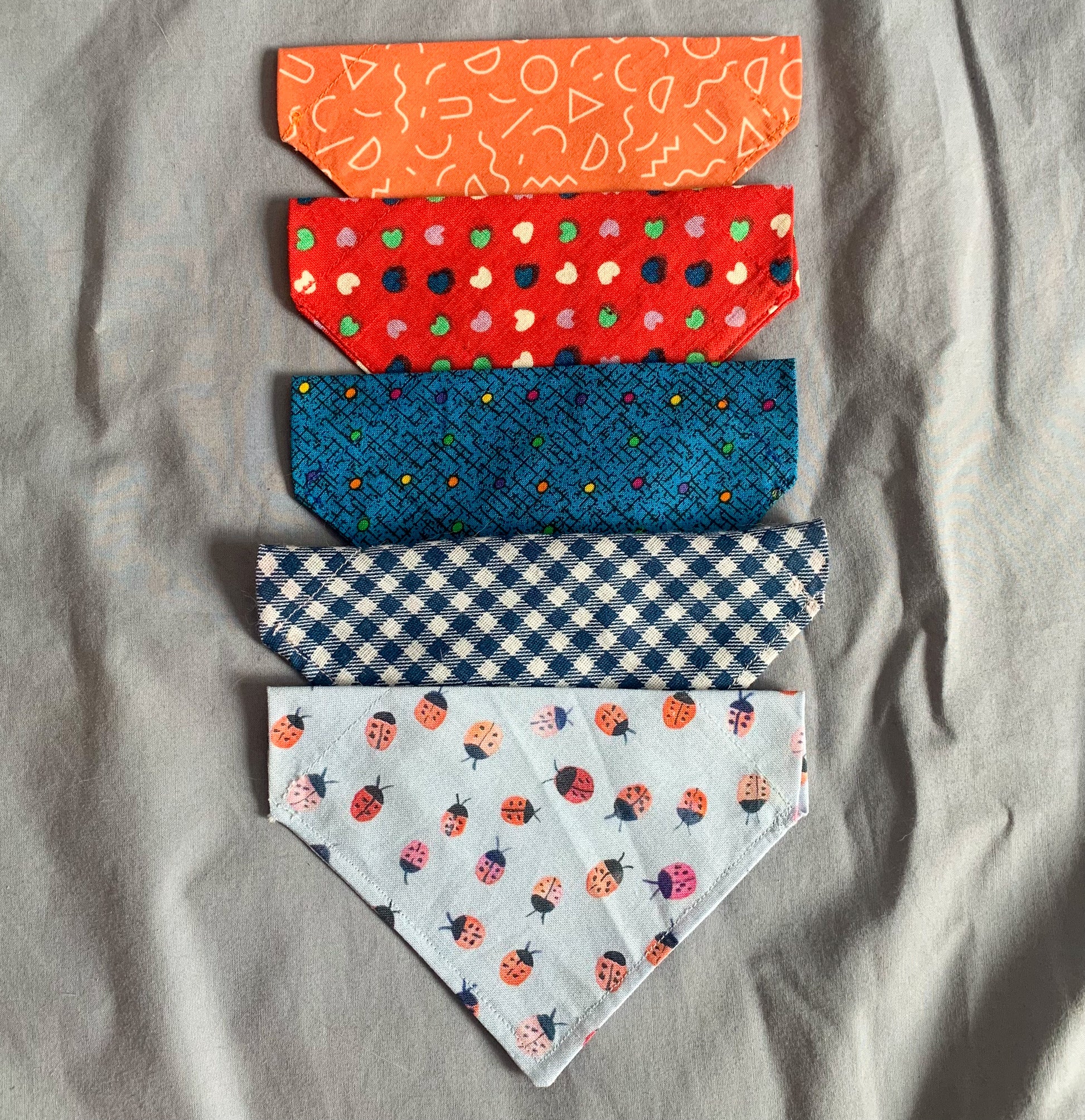 five triangle shaped cat bandanas in a line from top to bottom; orange, red heart print, blue dotted, blue gingham and blue ladybird polkadots