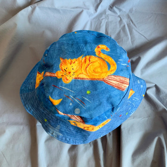 The top view of a blue bucket hat with an orange cartoon cat on a broom stick and stars on the top, on a blue background