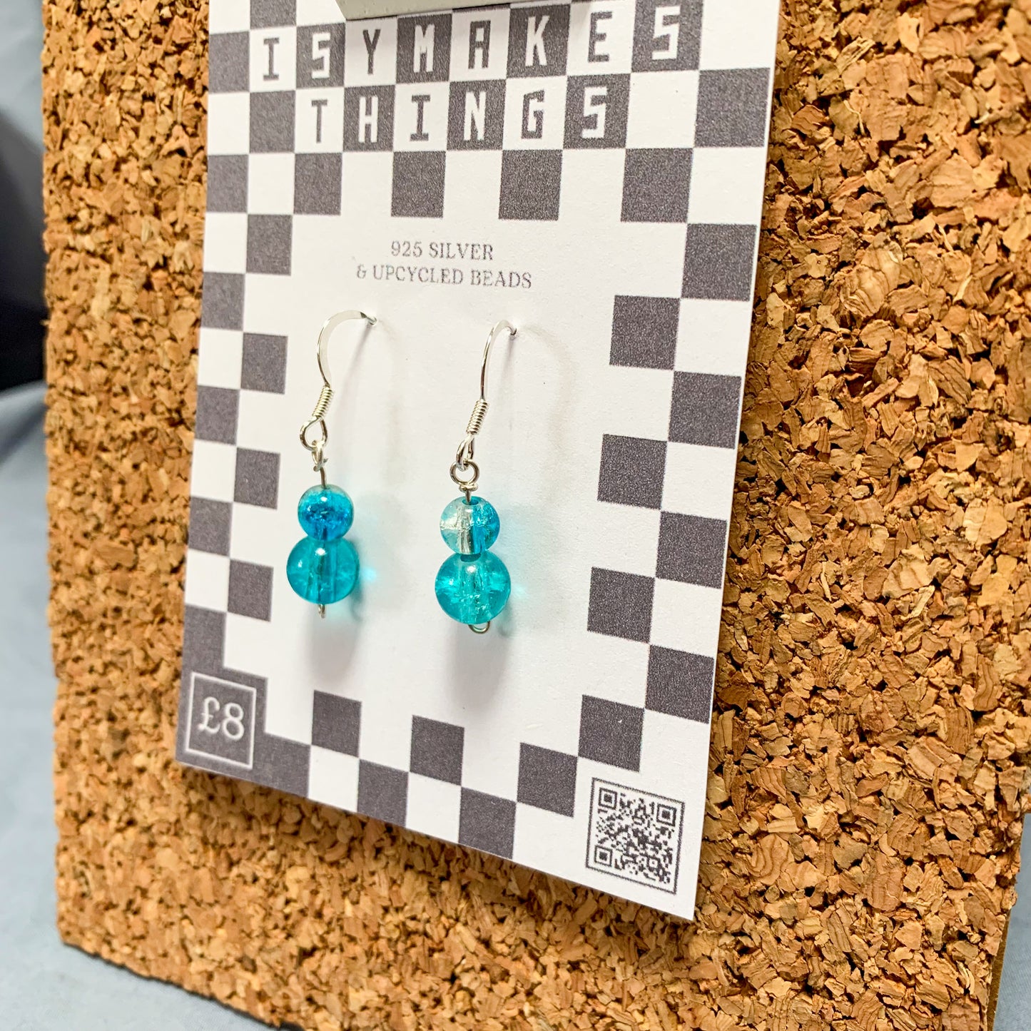 blue bead earrings on a checkerboard backing pegged to a corkboard against a blue background