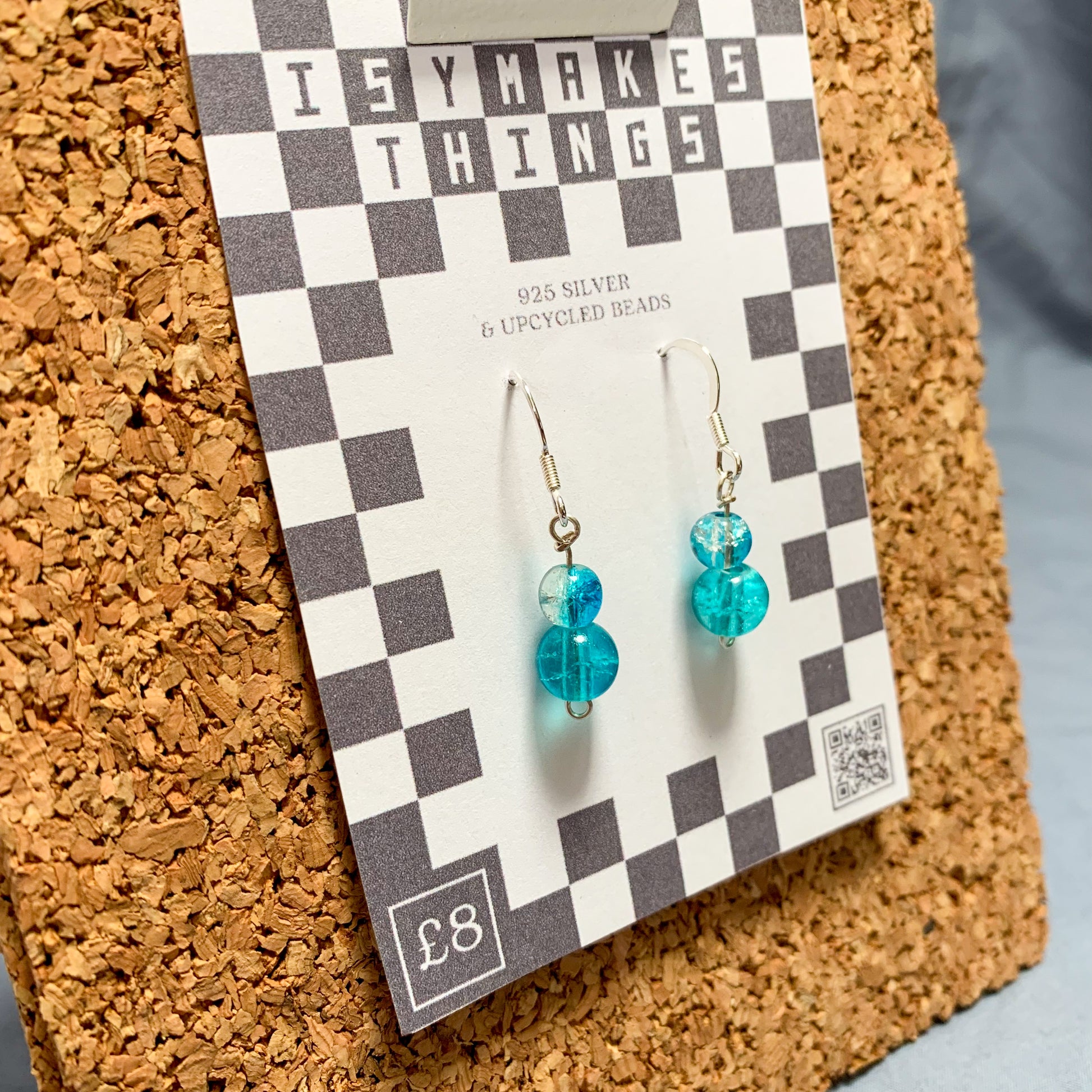 blue bead earrings on a checkerboard backing pegged to a corkboard against a blue background
