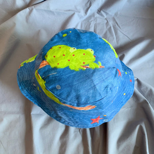 The top view of a blue bucket hat with a green cartoon frog on a broom stick and stars on the top, on a blue background