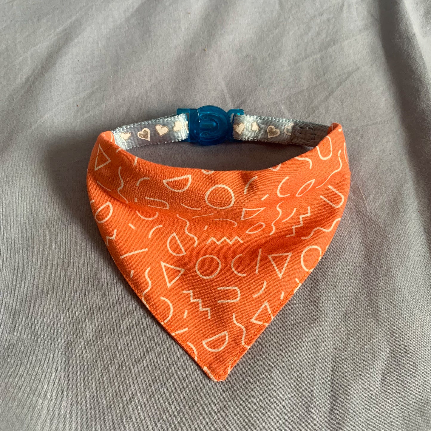 An orange abstract shape patterned triangle cat bandana on a blue collar sat on a blue background