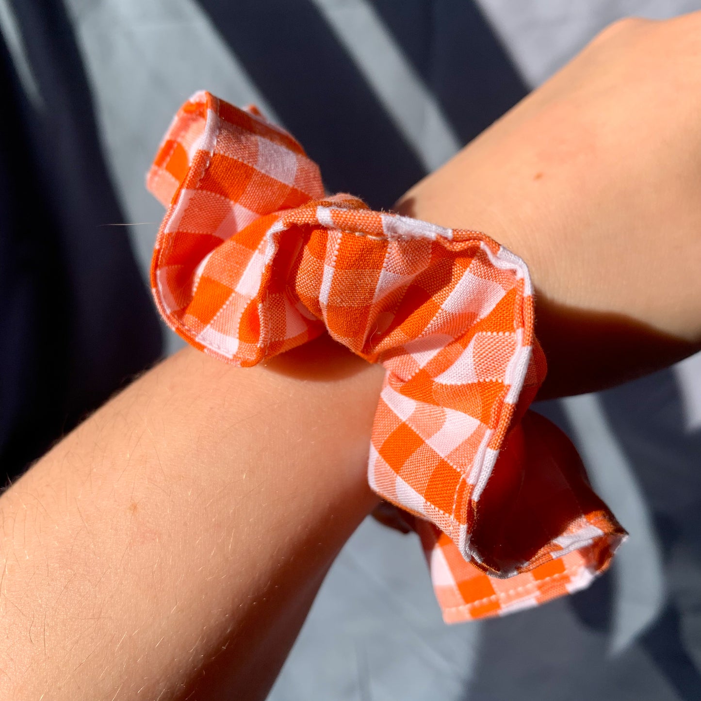 An orange gingham scrunchie showcased on a white persons wrist against a blue background