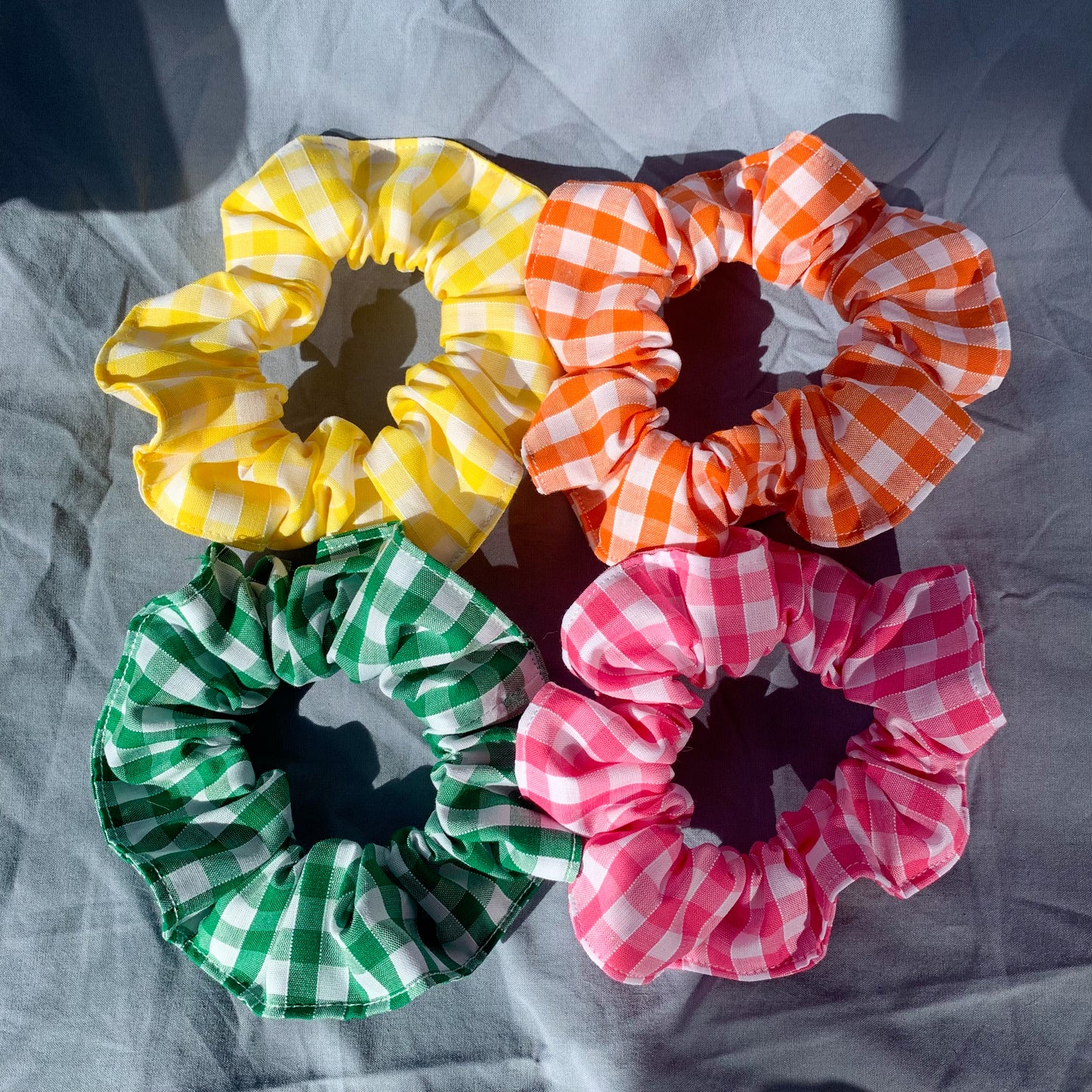 Four gingham scrunchies in a pile, yellow, organge, green & pink on a blue background