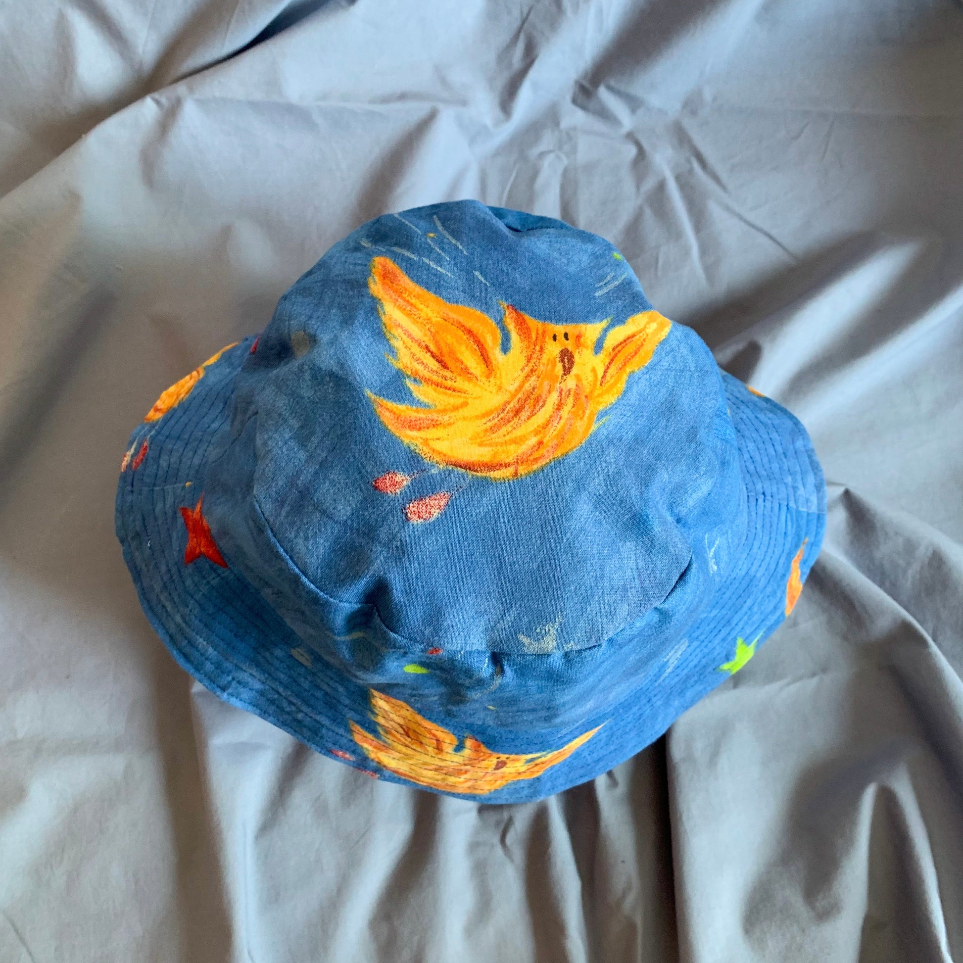 The top view of a blue bucket hat with an orange cartoon phoenix and stars on the top, on a blue background