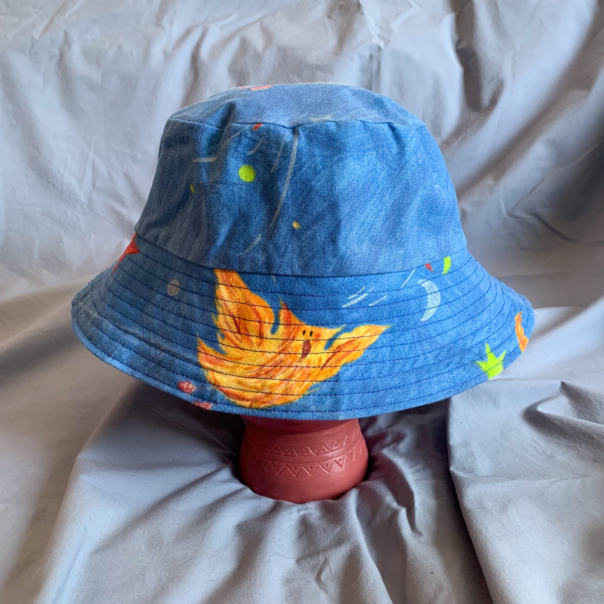 A blue bucket hat with part of a phoenix cartoon styled on a pink stone head against a blue background