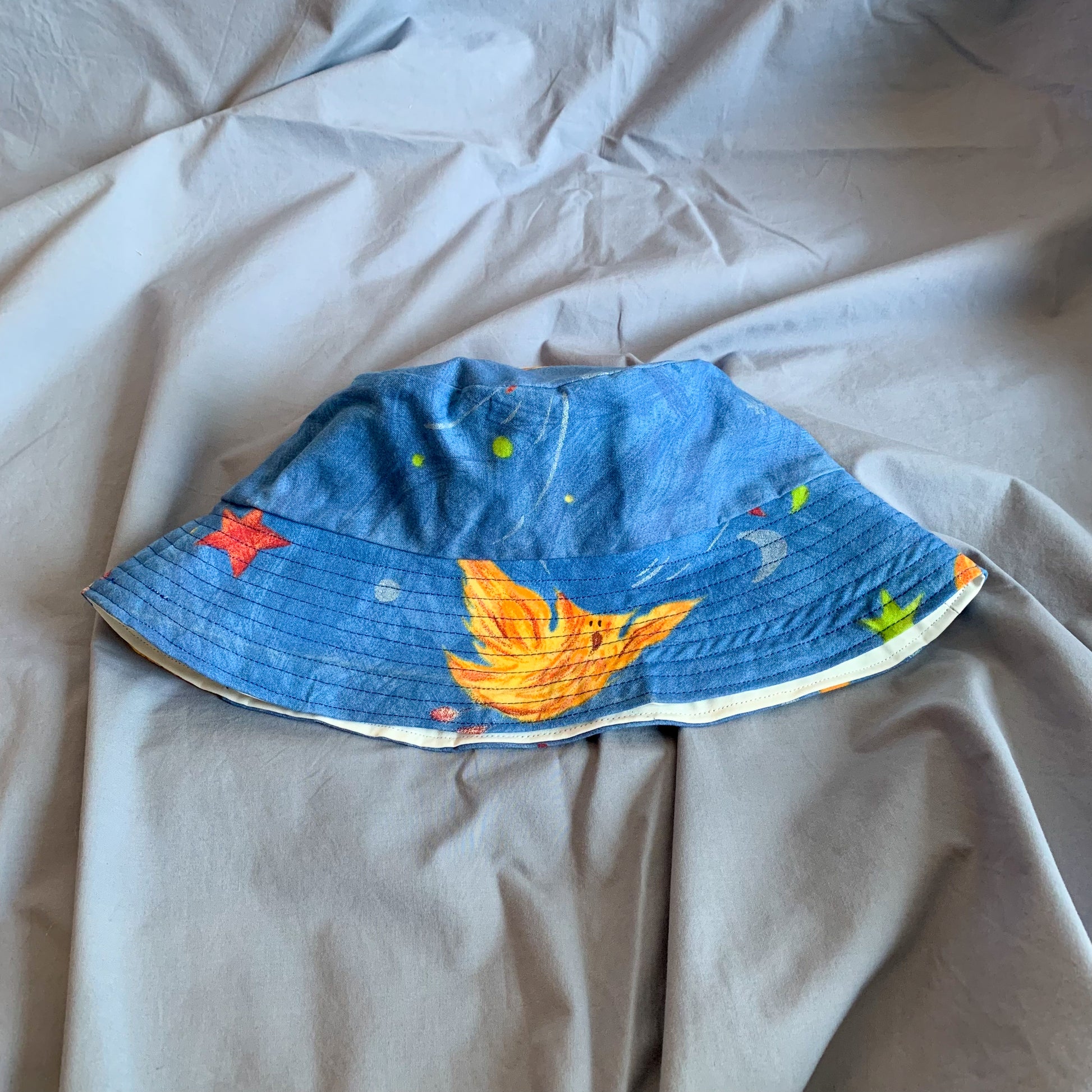 A blue bucket hat with part of a cartoon phoenix folded in half sat on a blue background