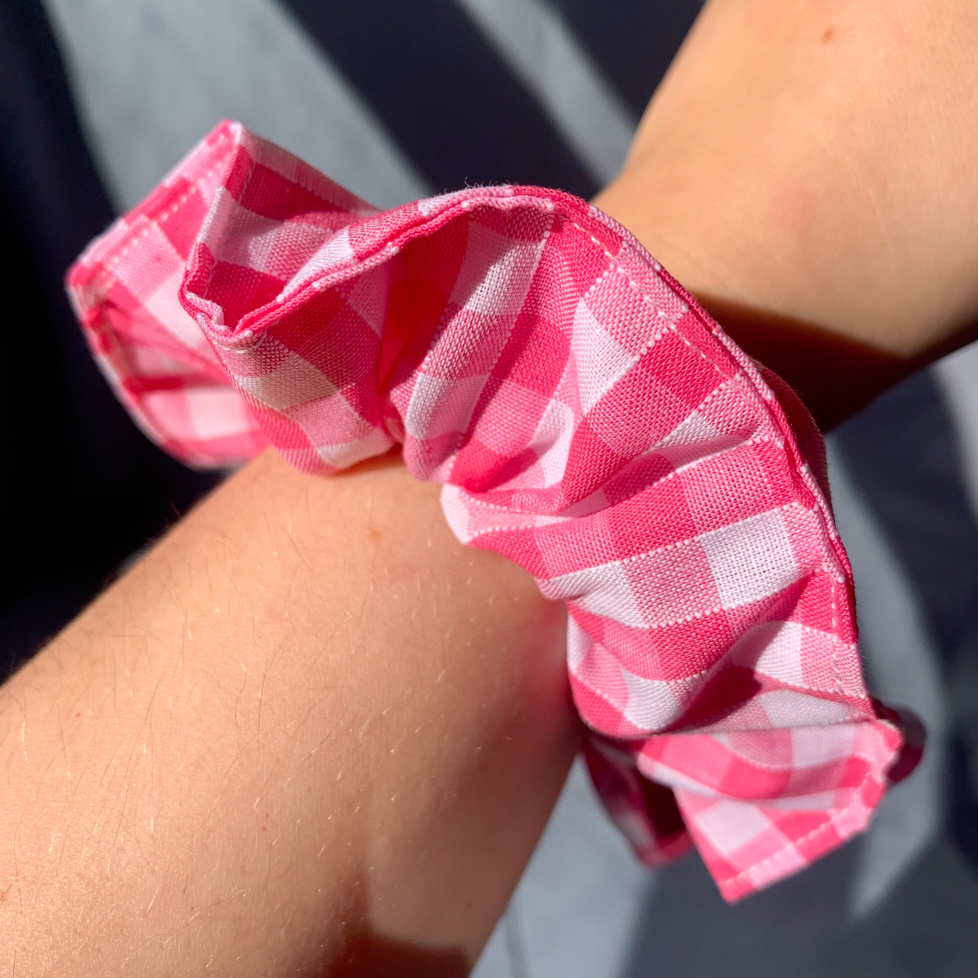 A pink gingham scrunchie showcased on a white persons wrist against a blue background