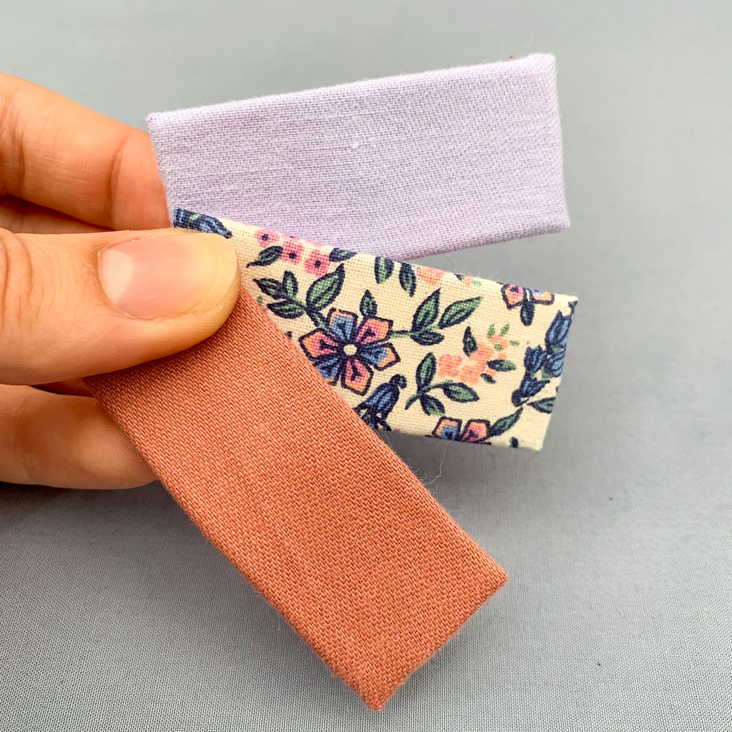 three pink & purple floral print hair clips held by a thumb fanned out across a blue grey background