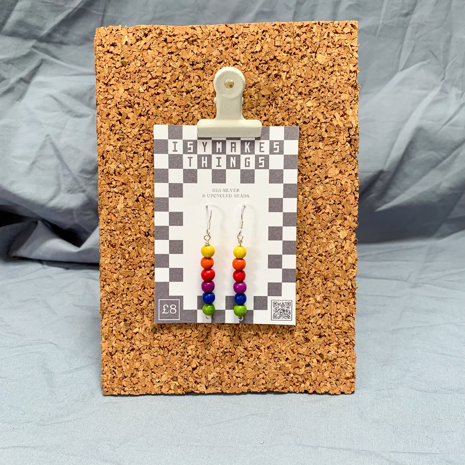Rainbow bead earrings on a checkerboard backing pegged to a corkboard against a blue background