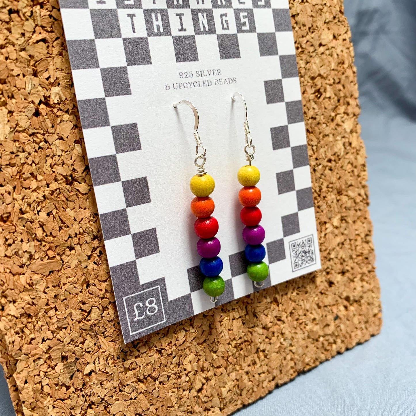 Rainbow bead earrings on a checkerboard backing pegged to a corkboard against a blue background