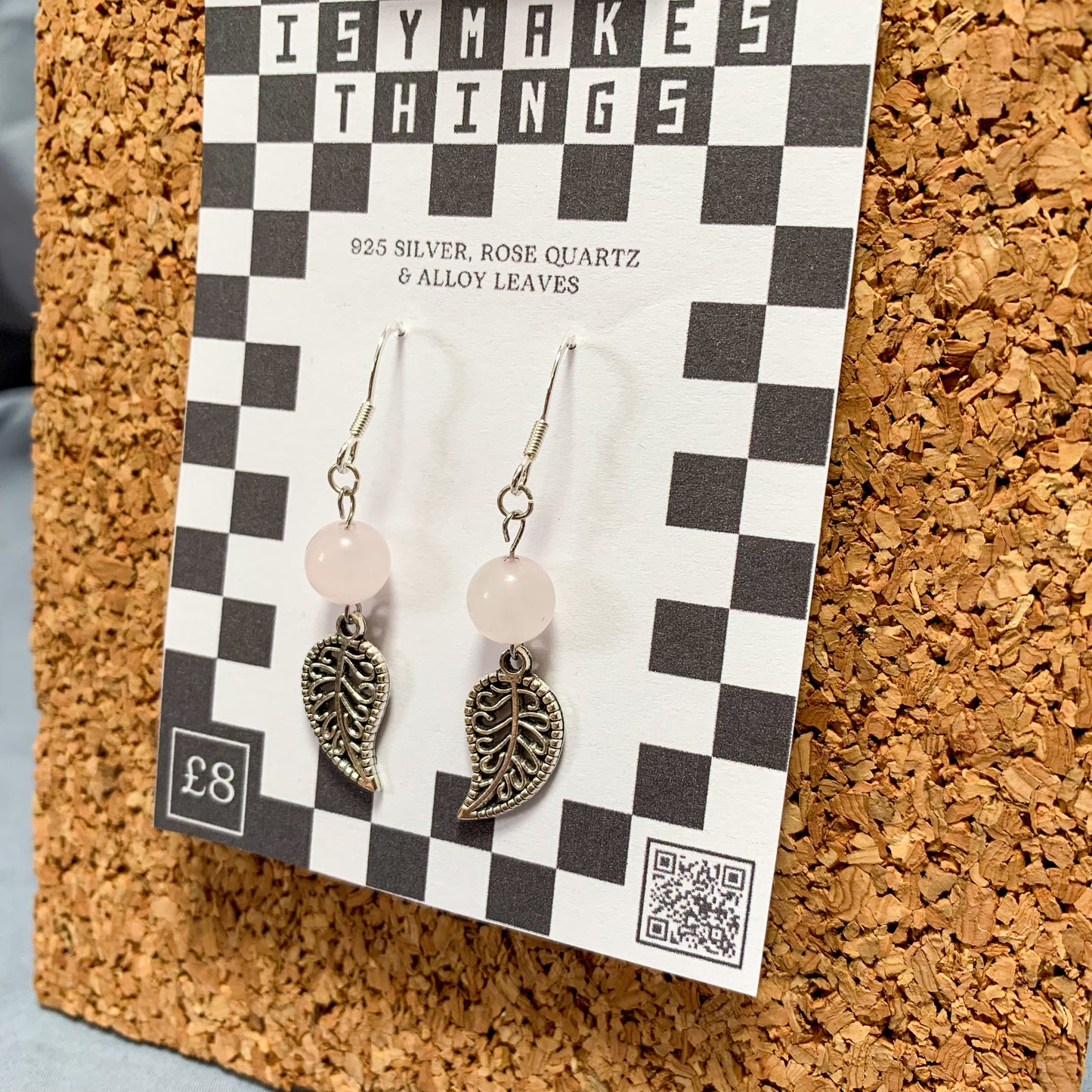 Rose quartz bead and leaf charm earrings on a checkerboard backing pegged to a corkboard against a blue background