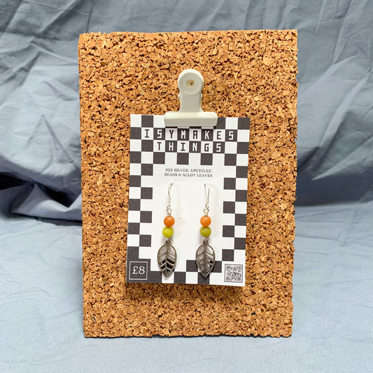 Orange & green leaf charm earrings on a checkerboard backing pegged to a corkboard against a blue background