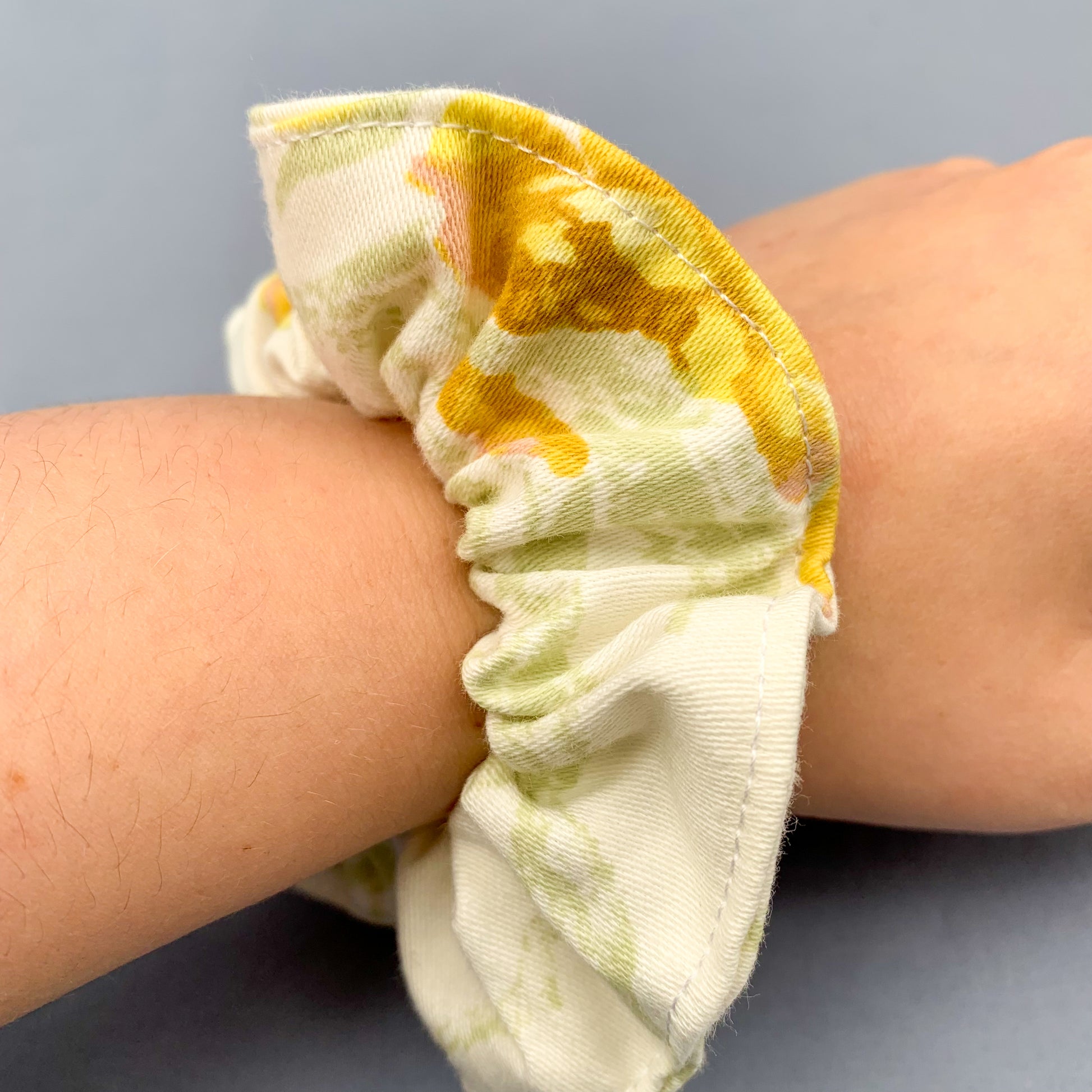 a white & yellow floral scrunchie on a white persons wrist against a blue background